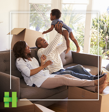 A couple laugh on the couch while playing with their toddler as unpacked moving boxes are stacked behind them.