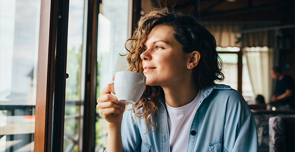 Girl looking out a window on a sunny day drinking a coffee.