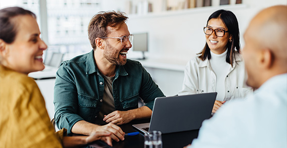 A group of coworkers chat and laugh in a meeting room.