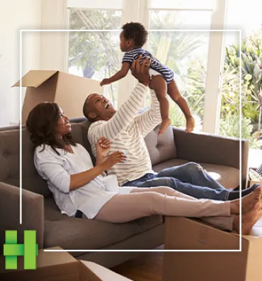 A couple laugh on the couch while playing with their toddler as unpacked moving boxes are stacked behind them.